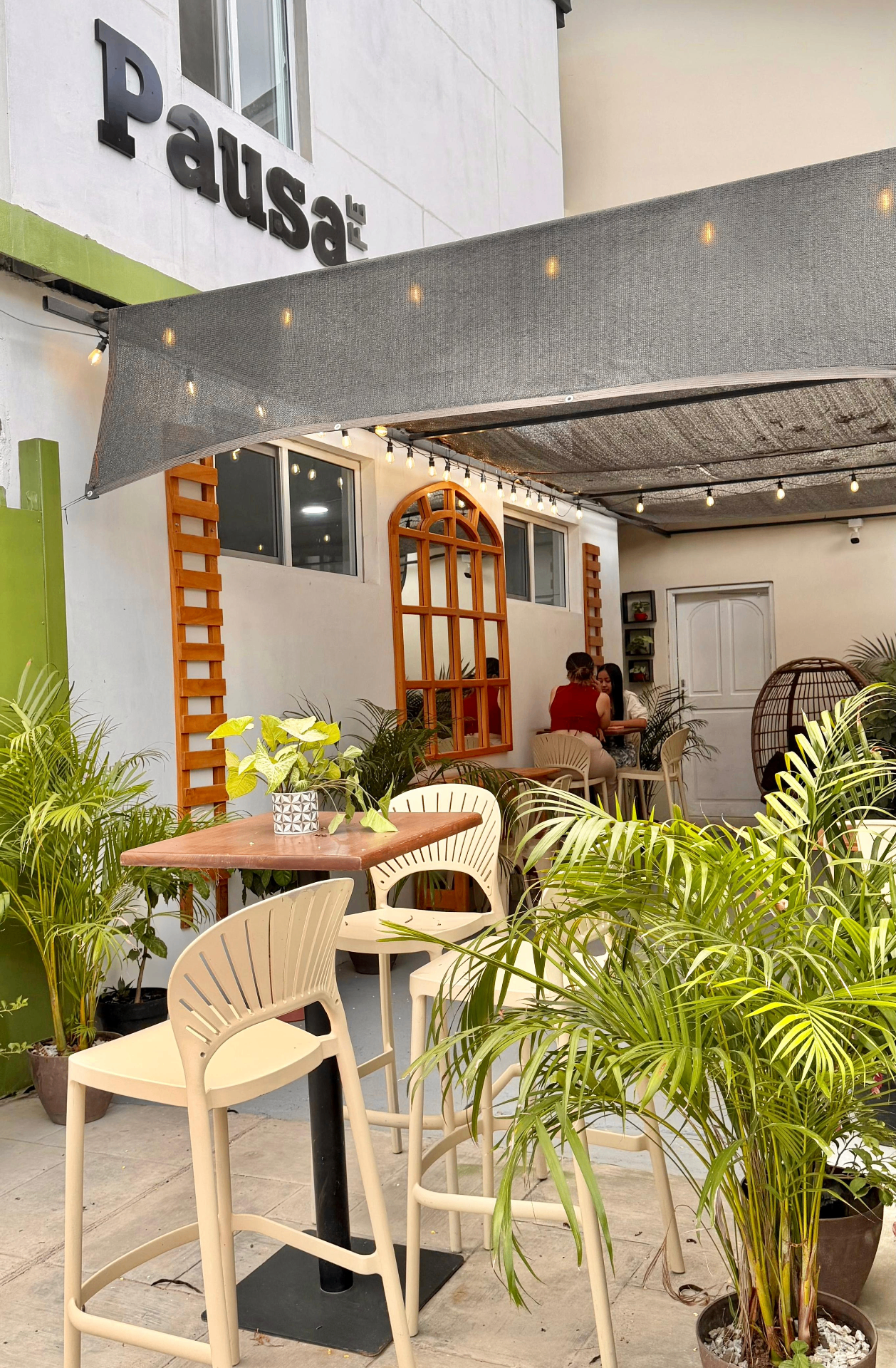 Terraza de Pausa Café con letrero, toldo, luces cálidas, plantas y mesas altas al aire libre